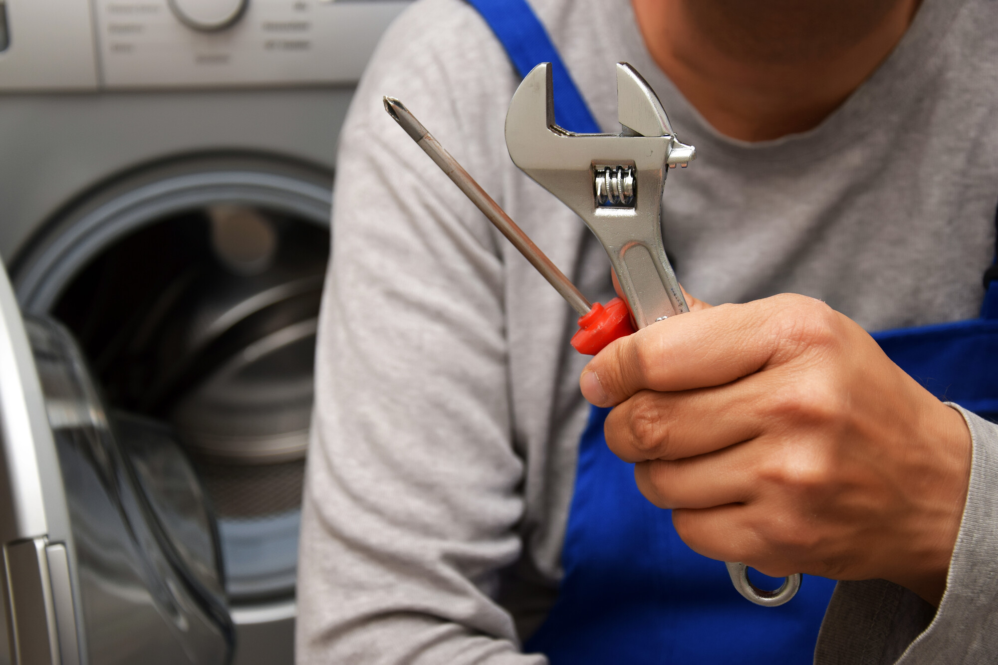 Tecnico in salopette blu e maglia grigia con chiave inglese e cacciavite rosso davanti a una lavatrice con oblò aperto